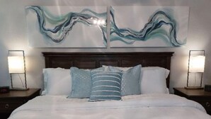 Una cama con ropa de cama blanca, dos mesitas de noche con lámparas, y un cuadro abstracto grande sobre la cama.
