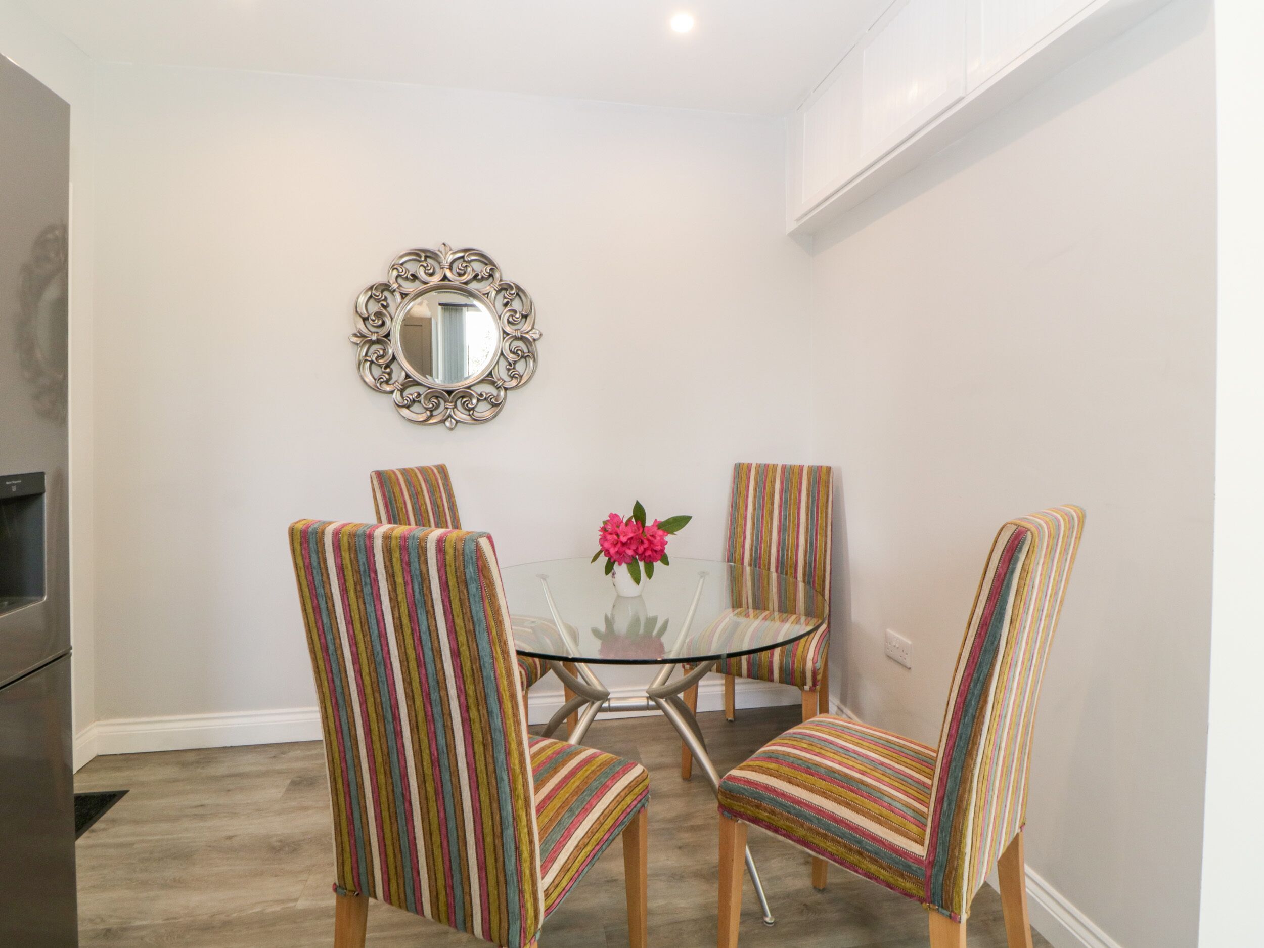 A dining area with a glass table surrounded by four striped chairs, a decorative mirror on the wall, and a vase with pink flowers.