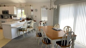A modern kitchen with white cabinetry, a central island, and a dining area with a wooden table and white chairs. - 4 bedroom accommodation in North Beach (North Beach)