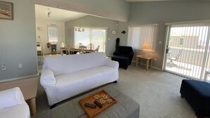 A living room with a white sofa, a dark armchair, a small table with a lamp, and a sliding glass door leading to an outdoor area. - 4 bedroom accommodation in North Beach (North Beach)