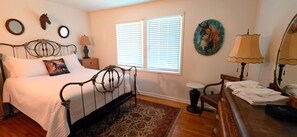 A bedroom with a bed, bedside tables, a nightstand, a chair, a dresser, a mirror, and a window with blinds. - Bostwick's Magnolia Manor (Wanchese)