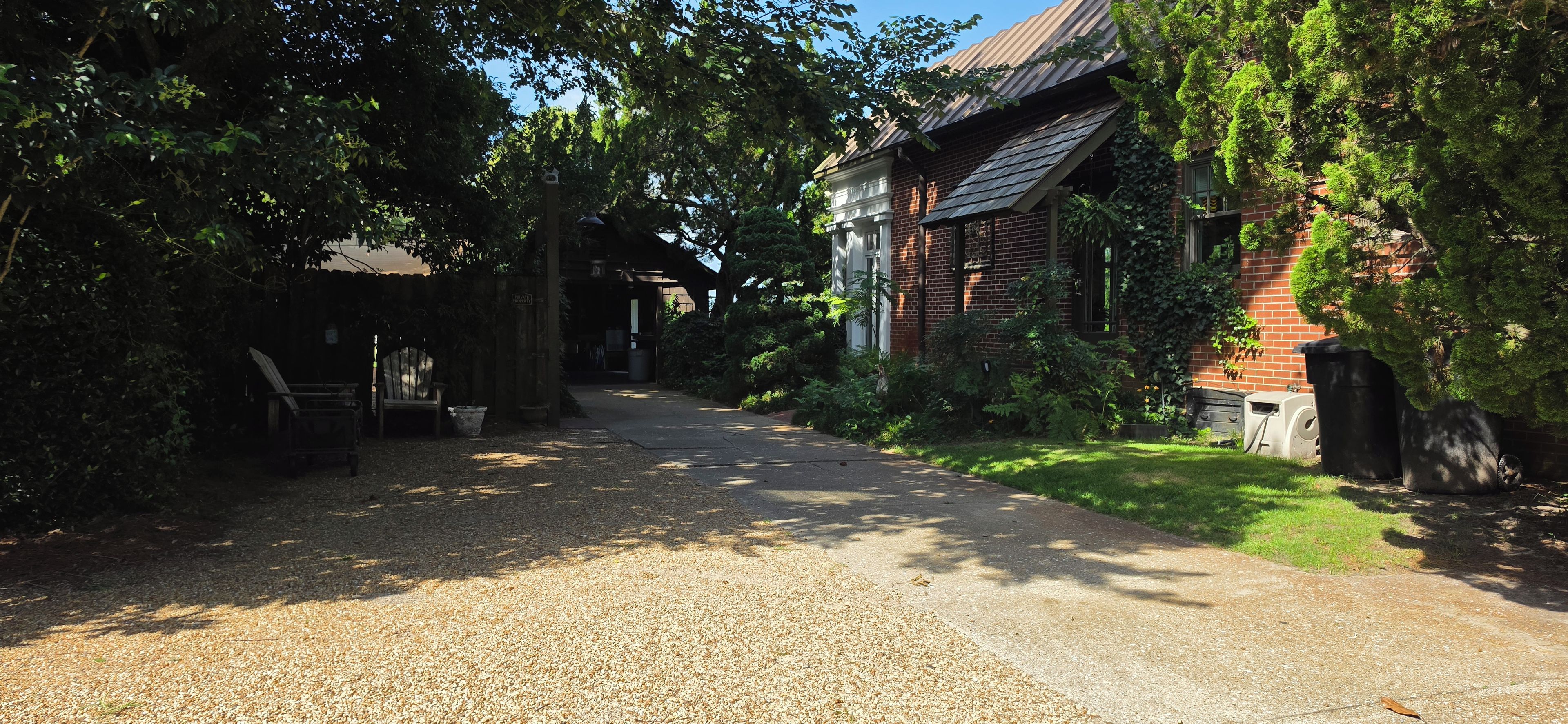 Une allée de gravier mène à un bâtiment en briques doté d’un porche abrité, le tout étant entouré d’une végétation luxuriante et d’arbres.