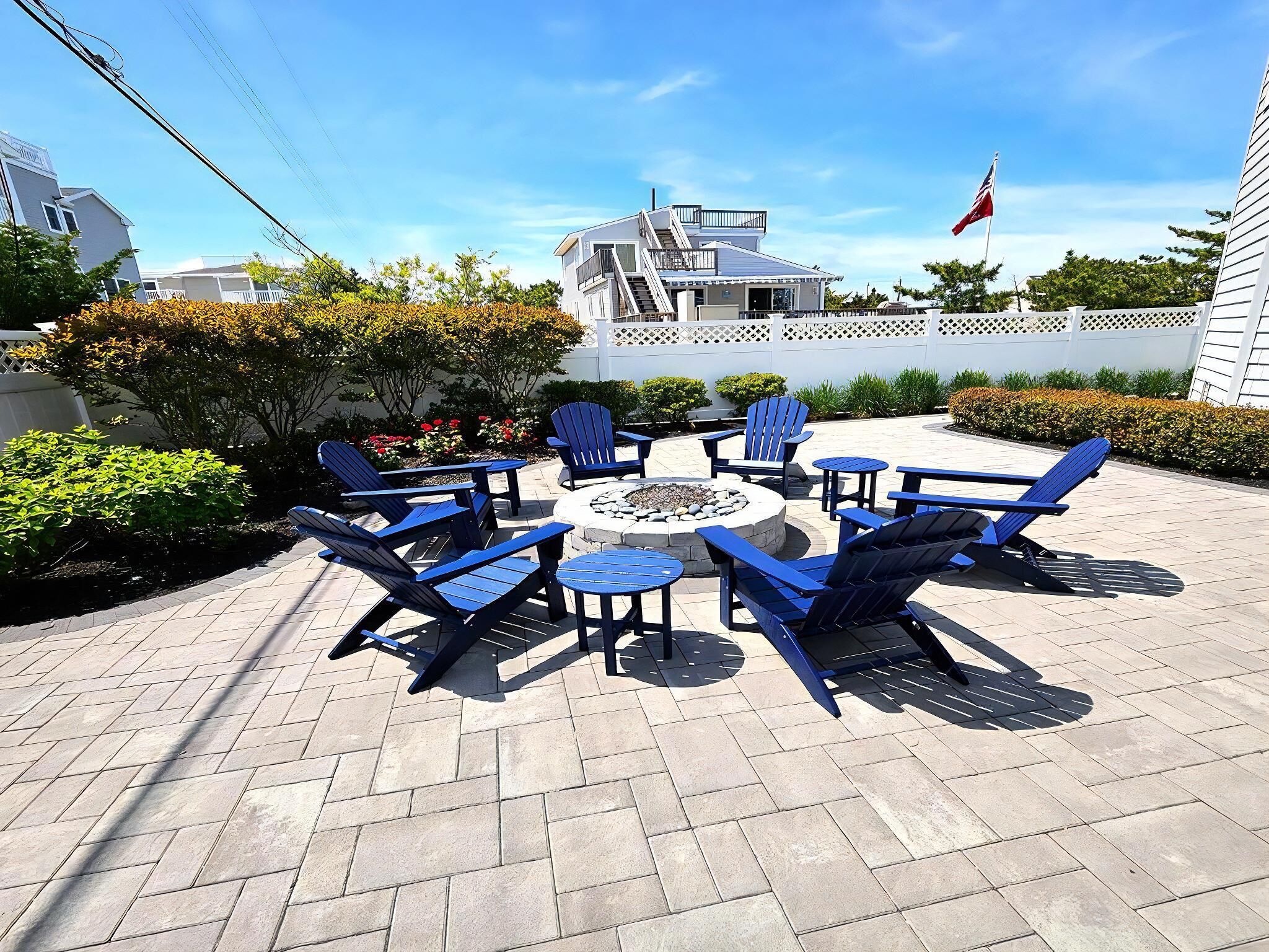 A patio with blue Adirondack chairs and a round table, surrounded by a white fence and greenery.