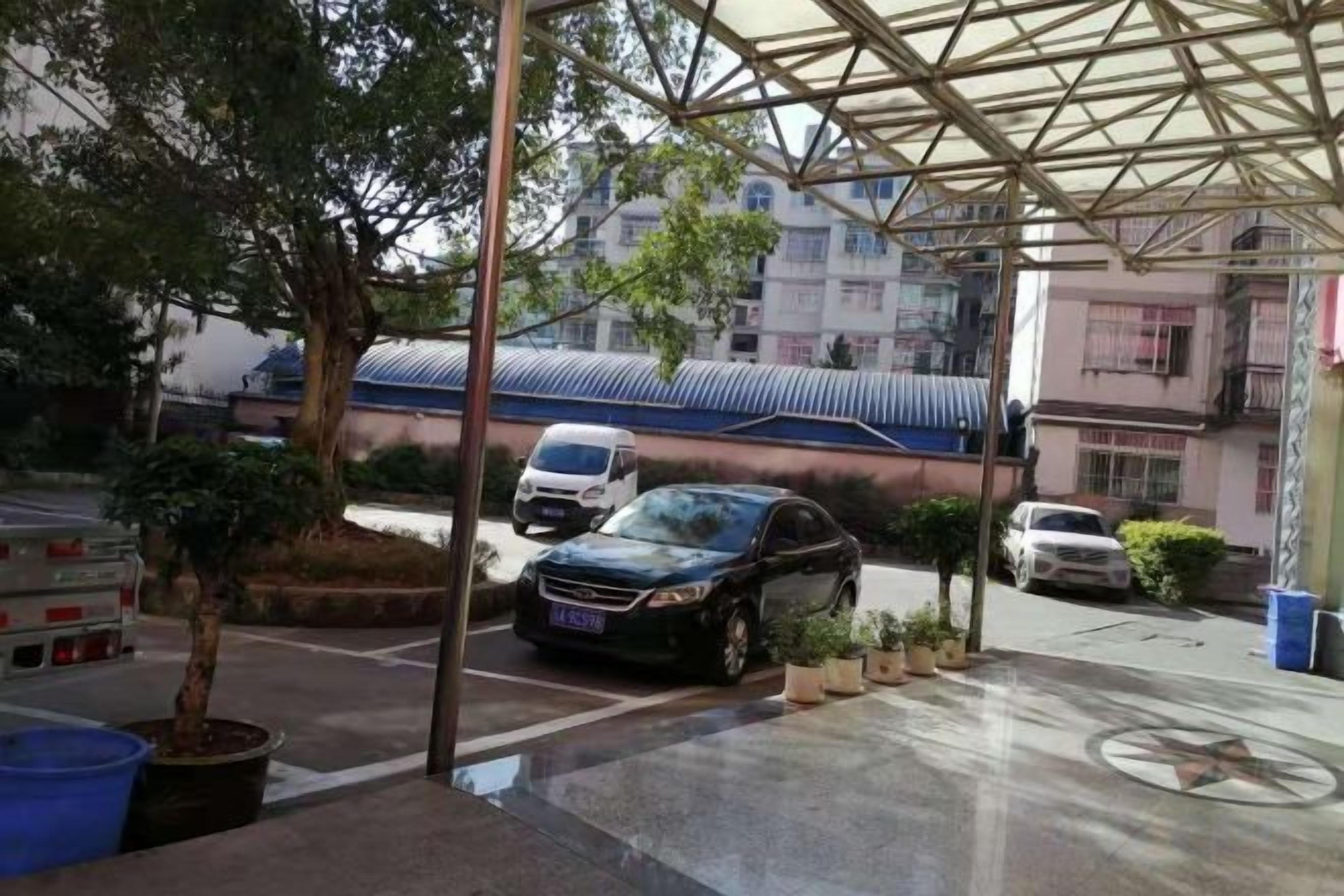 Un área de estacionamiento techada con coches, un árbol y un edificio con techo azul al fondo.