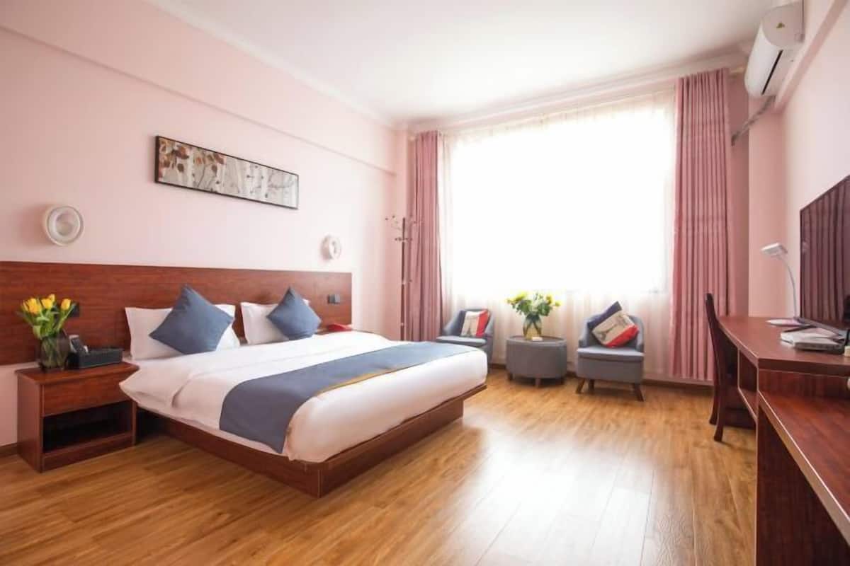 Une chambre d’hôtel avec un plancher en bois, un grand lit avec une literie bleue et blanche, un bureau avec une chaise, une petite table avec un vase de fleurs et une grande fenêtre avec des rideaux.