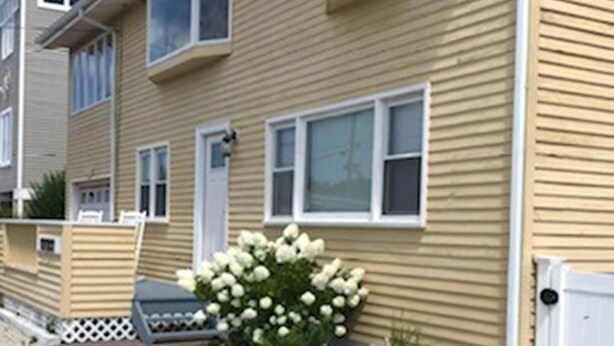 A two-story building with a beige exterior, white-framed windows, and a small garden with white flowers.