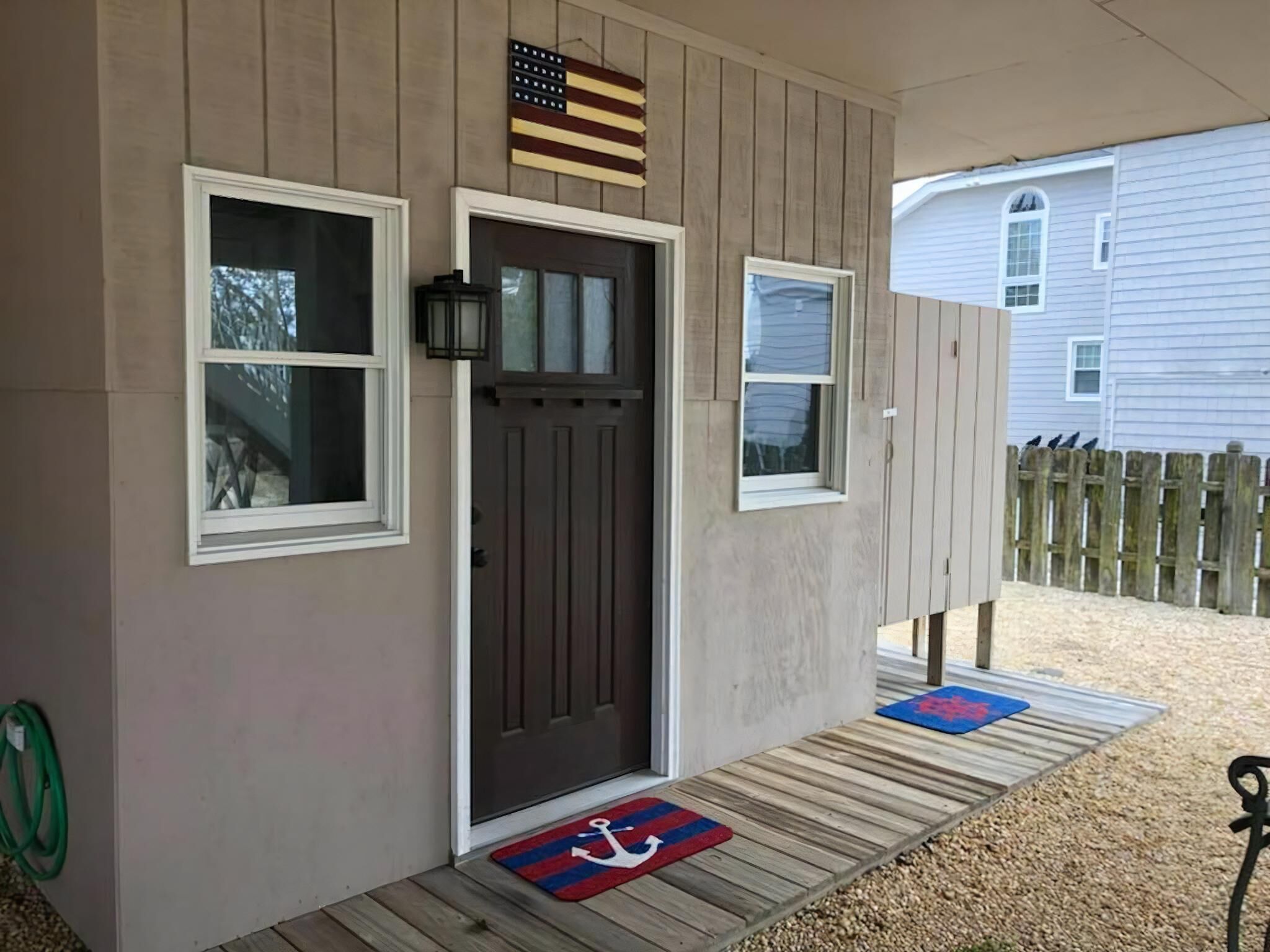 A house with a front porch, a wooden door, two windows, a small American flag, and a wooden deck with a rug.