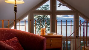 Une pièce avec un canapé rouge à motifs, un bureau en bois sur lequel se trouvent une tasse et un livre, et une vue sur un lac par de grandes fenêtres.