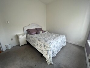 Quarto com uma cama de solteiro, mesa de cabeceira, lixeira pequena e uma parede lisa.