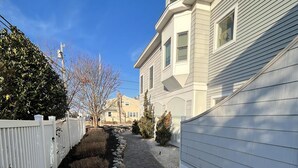 Une rue résidentielle avec un trottoir dallé, une clôture blanche et une rangée de maisons au revêtement clair.