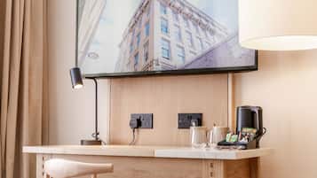 A hotel room with a wooden desk, a flat-screen TV mounted on the wall, a lamp, and a coffee maker.