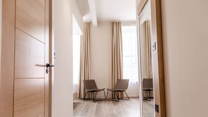 Une chambre d’hôtel avec une porte en bois, deux chaises, un grand miroir et une vue sur l’extérieur à travers des rideaux transparents.