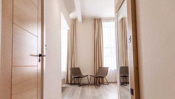 Hotelkamer met een houten deur, twee stoelen, een grote spiegel en uitzicht buiten door dunne gordijnen.