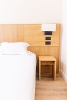 Una cama bien hecha con sábanas blancas, cabecera de madera, una mesita de noche de madera y un panel de control fijado a la pared.