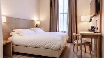 A hotel room with a bed, bedside table, chair, desk, lamp, and a TV.