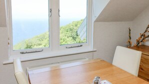 Una habitación con una mesa de madera, sillas blancas, vista al mar y una ventana con persiana de madera.