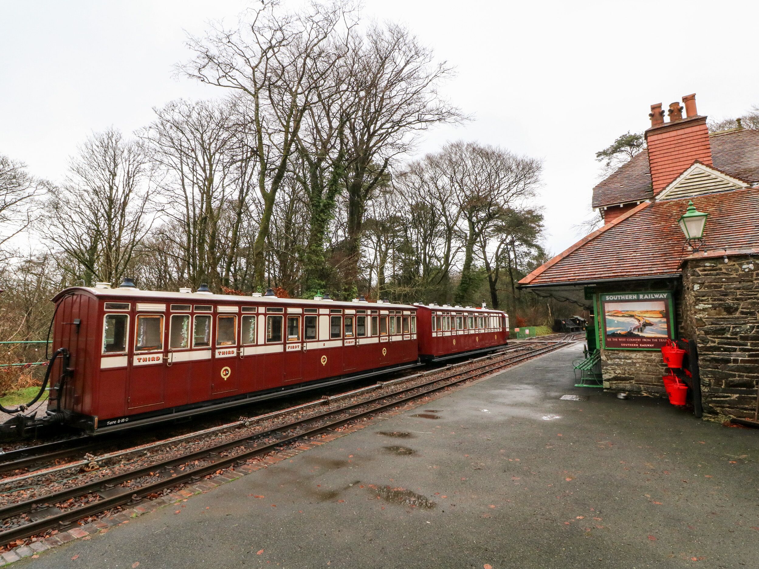 A vintage train at a station platform with a stone building and trees in the background.