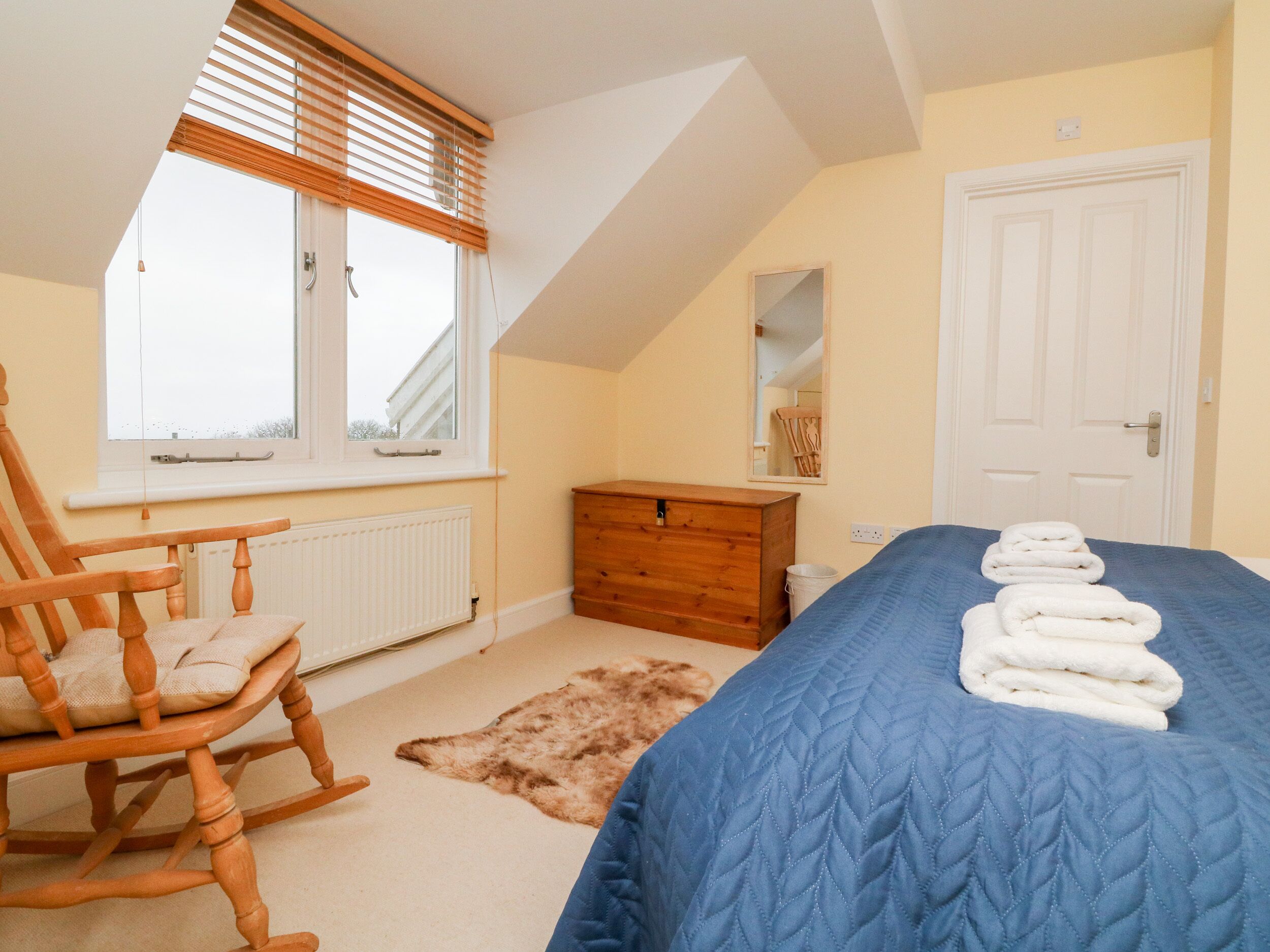 A bedroom with a sloped ceiling, a wooden rocking chair, a dresser, a bed with folded towels, a fur rug, and a window with blinds.