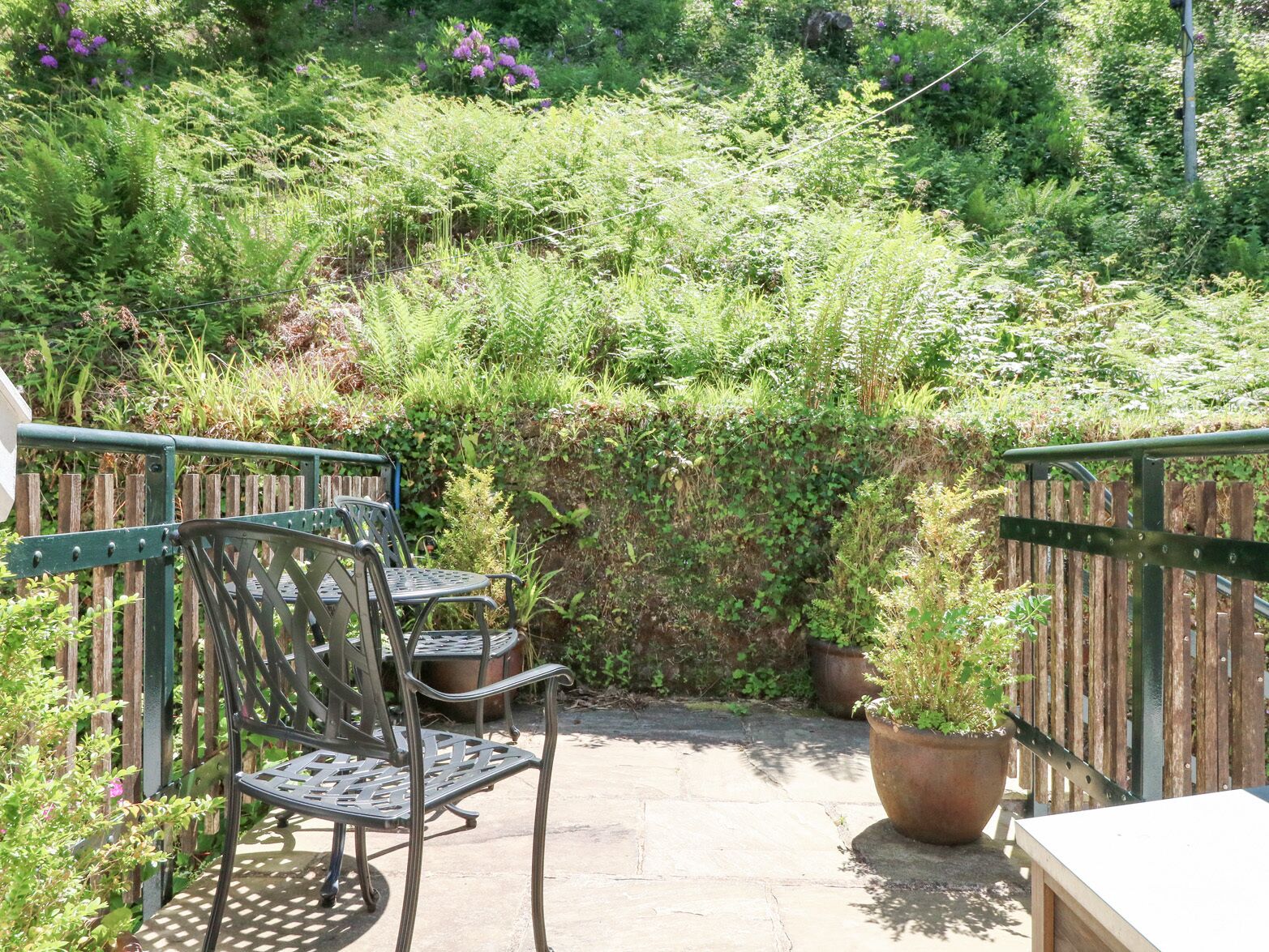 A patio with metal chairs and a table, surrounded by lush greenery and a wooden fence.