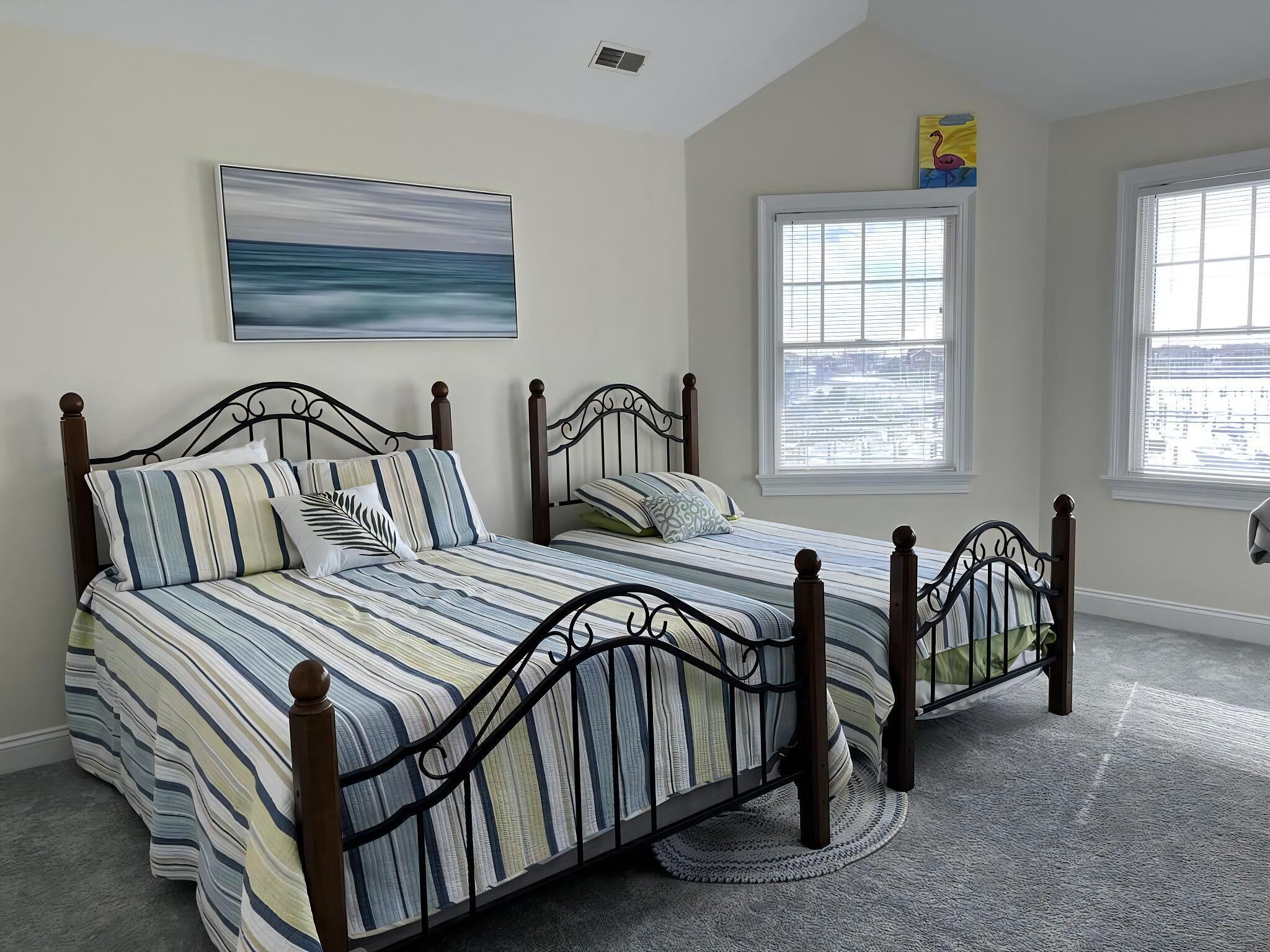 A bedroom with a double bed, a window with blinds, a painting on the wall, and a carpeted floor.