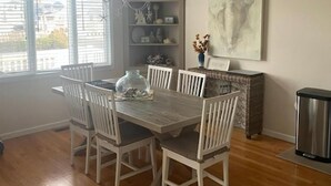 Un coin repas comprenant une table en bois, des chaises blanches, un lustre, une fenêtre avec des stores et un élément décoratif mural.