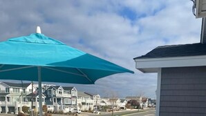 Une terrasse avec des chaises blanches et une table, un parasol bleu, et des maisons en arrière-plan.