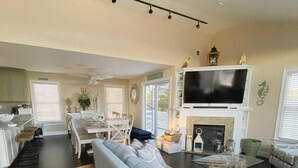 A spacious living room with a large sofa, a dining area with a table and chairs, a fireplace, and a television mounted on a white mantelpiece. - Ocean views- luxurious vacation rental on LBI (Brant Beach)