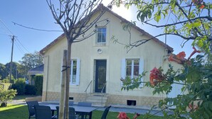 A two-story house with a front porch, a dining area with outdoor furniture, and a garden with flowering plants.