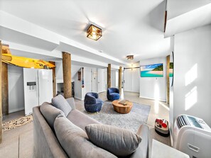 Ein modernes Wohnzimmer mit einer Couch, zwei Sesseln, einem Couchtisch und einem Fernseher. Holzsäulen, ein Kühlschrank und ein großes Fenster sind ebenfalls vorhanden.