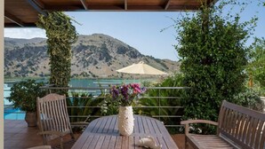 Een houten terras met een tafel, stoelen en een bloevas. Een grote parasol is op de achtergrond zichtbaar, en er is een bergachtig landschap te zien.