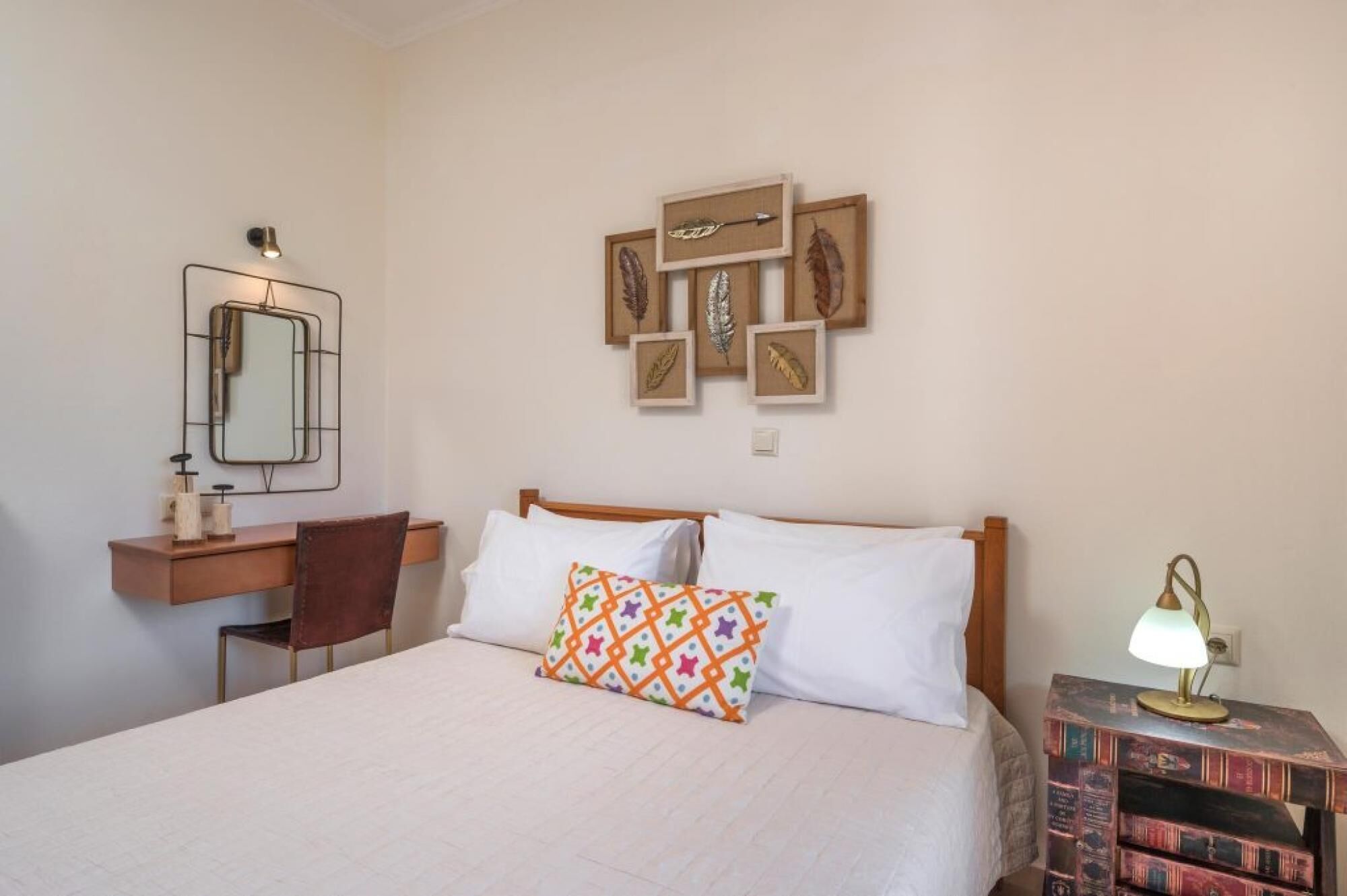 A bedroom with a bed, a bedside table with a lamp, a chair, a desk with a mirror, and wall-mounted artwork.