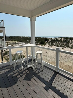 A wooden deck with white railings, two white stools, and a clear view of the beach and dunes.