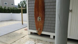 Ein Surfbrett aus Holz an einem weißen Gebäude mit grauer Fassade, einem weißen Zaun und einer Schotterterrasse mit trittfesten Steinen.
