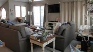 Ein Wohnzimmer mit Deckenventilator, Flachbildfernseher und einem großen Fenster mit Vorhängen.