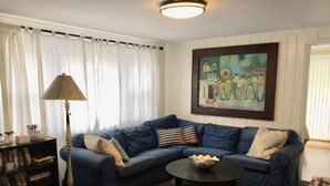 Ein Wohnzimmer mit einem dunkelblauen Ecksofa, einem runden Couchtisch, einer Lampe, einem Bücherregal und einem Gemälde an der Wand.