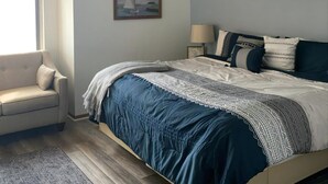 Una camera da letto con un letto, una ventola a soffitto, una finestra con tende, una sedia e quadri appesi al muro.