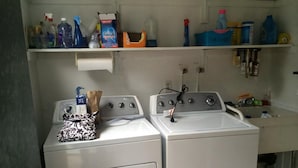 A laundry area with two white washing machines, a shelf with cleaning supplies, and a countertop with a sink. - Outstanding Bayviews from this 1st floor duplex (Surf City)