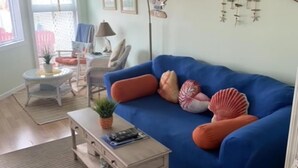 Un salon avec un canapé bleu, une petite table basse et une plante en pot. On aperçoit une chaise et une lampe au fond de la pièce.