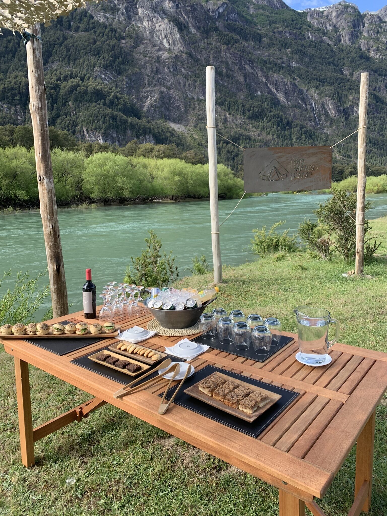 Une table en bois installée à l’extérieur, avec de la nourriture, des boissons et une bouteille de vin, d’où l’on peut admirer une rivière et des montagnes.
