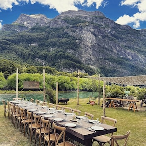 A long dining table set outdoors with chairs, a signboard, and a fire pit, surrounded by a mountainous landscape and a river. - Rio Manso Camp - Glamping Bariloche (San Carlos de Bariloche)