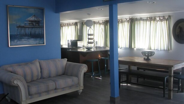 Een woonkamer met een bank, een eettafel met stoelen, een keukenhoek, en een bordje met de tekst 'BEACH HOUSE'.