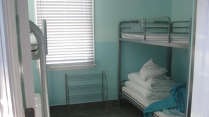 Een kleine eenpersoonskamer met een metalen stapelbed, een raam met zonwering, een metalen radiator en een gestreept tapijt op de vloer.