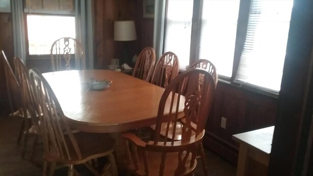A dining area with a wooden table and chairs, a lamp, and windows with blinds. - 3rd FROM THE OCEAN - SLEEPS 14! (Barnegat Light)