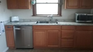 A kitchen with wooden cabinets, a stainless steel dishwasher, a microwave, and a window above the sink. - 3rd FROM THE OCEAN - SLEEPS 14! (Barnegat Light)