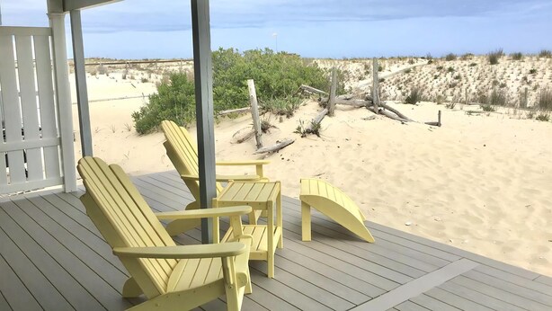 A covered outdoor seating area with wooden chairs and a table, overlooking a sandy beach with dunes and sparse vegetation. - Pet friendly beachfront vacation rental on LBI (Brant Beach)