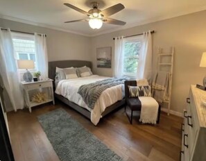 Ein Schlafzimmer mit Bett, Nachttischen, Schreibtisch, Stuhl, Stehlampe, Deckenventilator und einem Fenster mit Vorhängen.