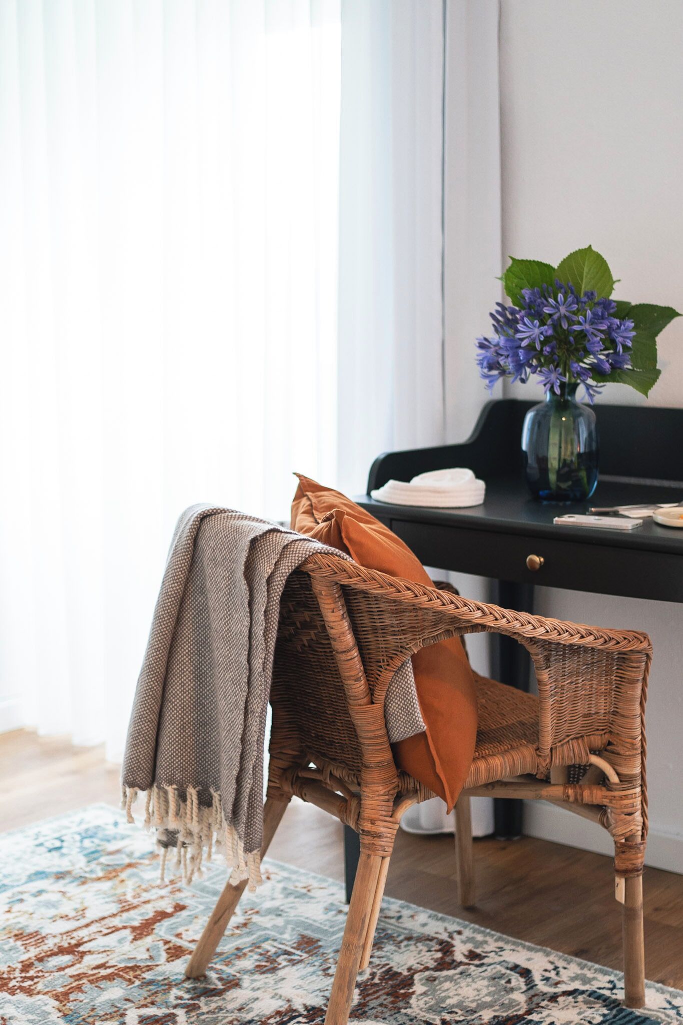 A wicker chair with a blanket and a cushion, a desk with a vase of flowers, and a patterned rug.