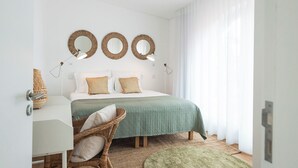 A bedroom with a bed, two wall-mounted mirrors, a bedside table, a wicker chair, and a green rug. - Azores Green Villas (Ponta Delgada)