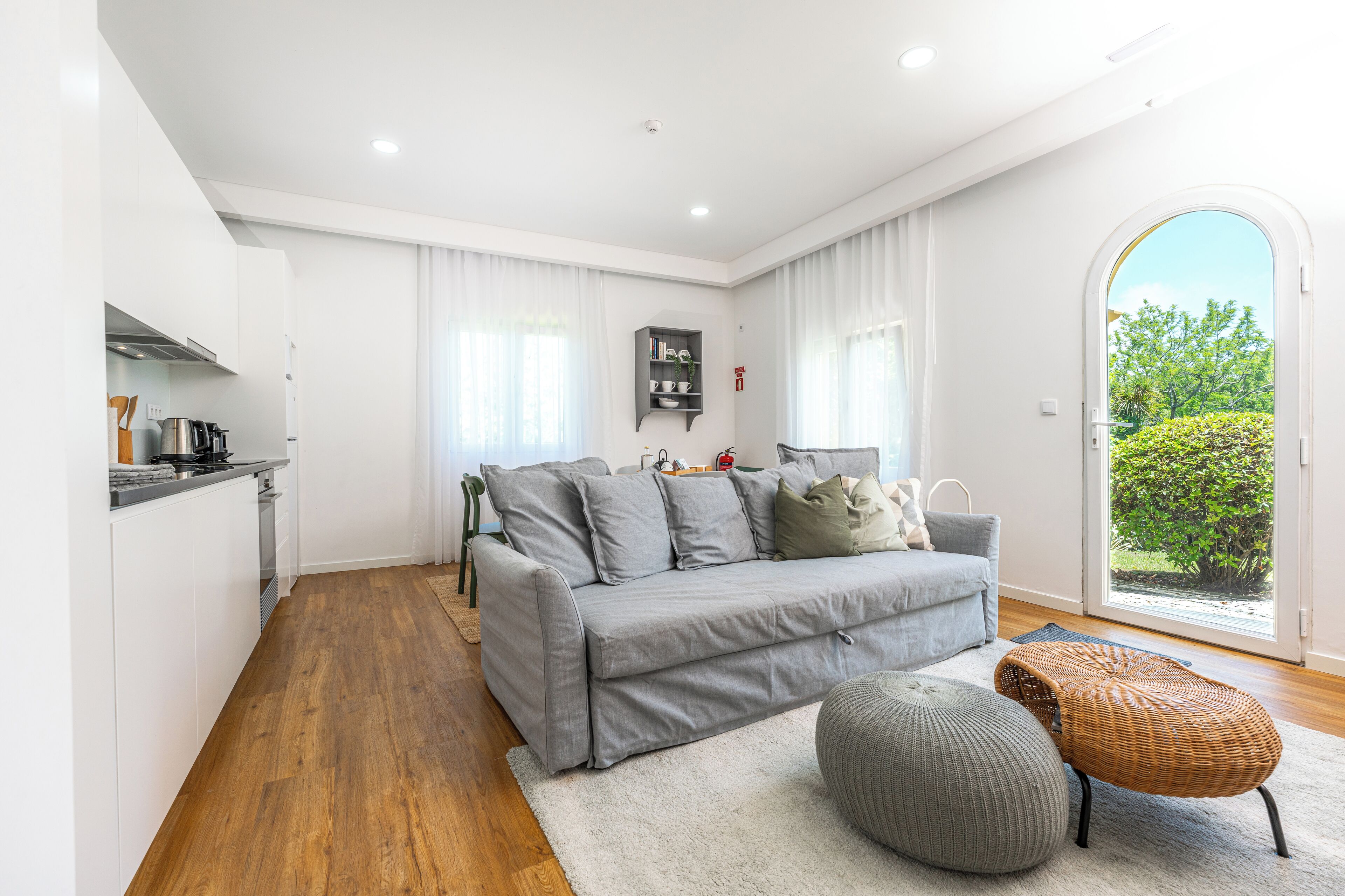 A modern living room with a grey sofa, a small table, and a wicker ottoman. There is a kitchen area with white cabinets and a wooden floor.