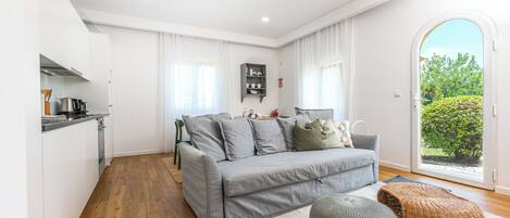 A modern living room with a grey sofa, a small table, and a wicker ottoman. There is a kitchen area with white cabinets and a wooden floor.